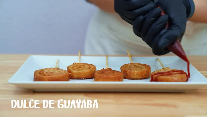 Colocar los rollitos de pan en la mezcla de mantequilla, azúcar y agua, y coronar con dulce de guayaba derretido y queso crema (opcional).
