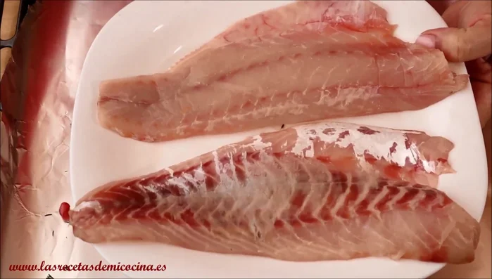 Coloca la lubina sobre la granada: Pon la lubina encima de la granada en la bandeja. Añade eneldo, pimienta, más granada y un chorrito de aceite de oliva y vino blanco.