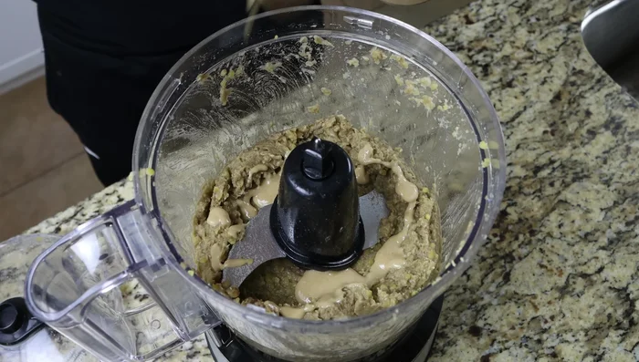 Agregar la pasta de tahini (3 cucharadas, 45g), ajo (2 dientes), comino molido (1/2 cucharadita, 1.5g), pimentón (1/4 cucharadita, 0.5g+), aceite de oliva virgen extra (2 cucharadas, 30ml), jugo de limón (1 cucharada, 15ml), sal y pimienta al procesador de alimentos. Procesar durante 1-2 minutos hasta que esté bien mezclado.