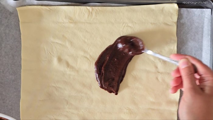 Extiende la plancha de hojaldre y coloca el chocolate derretido, los bizcochos de soletilla empapados en leche y el plátano en láminas encima. (11.4, 18.279, 28, 31.279, 46.36)
